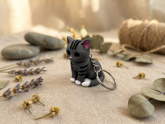 3D-gedruckter Katzen-Schlüsselanhänger grau-getigert mit weißen Pfoten und Metallring, auf Leinenstoff mit Trockenblumen, von fks-design
